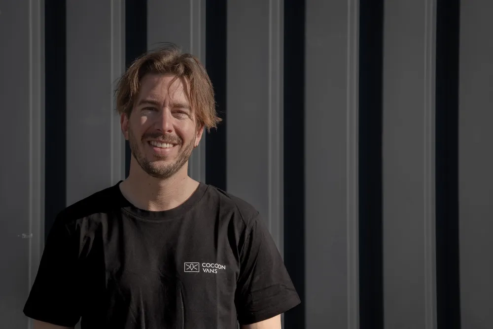 Leo lächelt in die Kamera und steht vor einer dunklen Wand aus Wellbleck