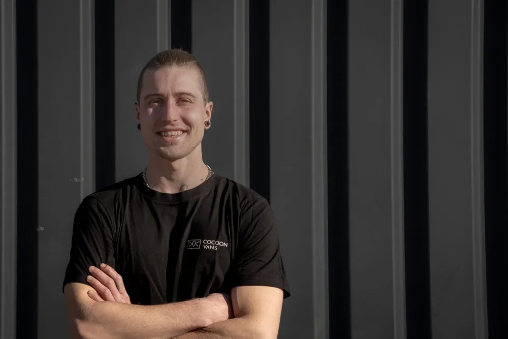 Ivo steht vor einer Antrazit schwarzen Wand und blickt in die Sonne