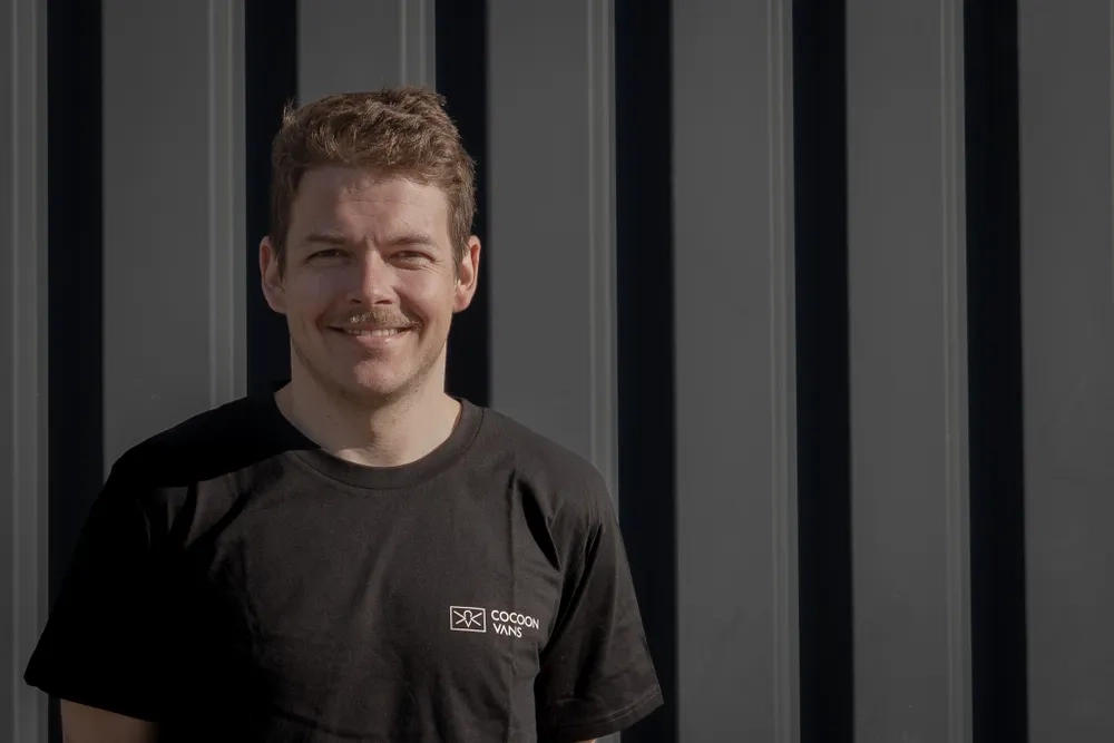 Jonas trägt ein dunkles T-Shirt mit dem Cocoon Vans Logo und lächelt in die Kamera. Er steht vor einer dunklen Wand im freien